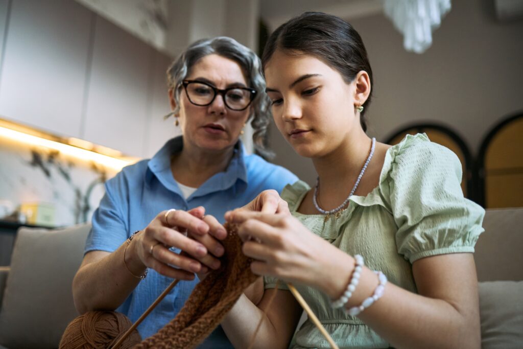 Teenage girl learning knitting from senior woman at home, practicing handmade craft skills across generations.