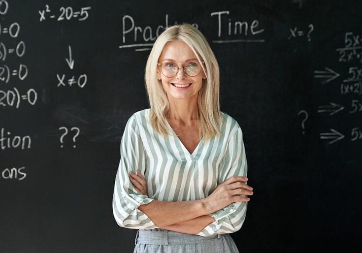 Happy smiling middle aged woman mature high school education math teacher or college professor wearing glasses standing at blackboard background. Confident female professional educator portrait.